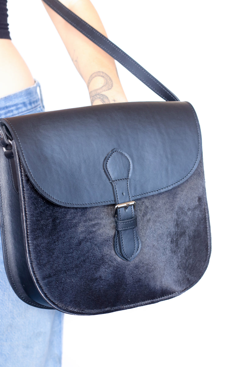 RoHo's handmade crossbody saddle purse in black hide and finished leather being held against a white back ground by a model 